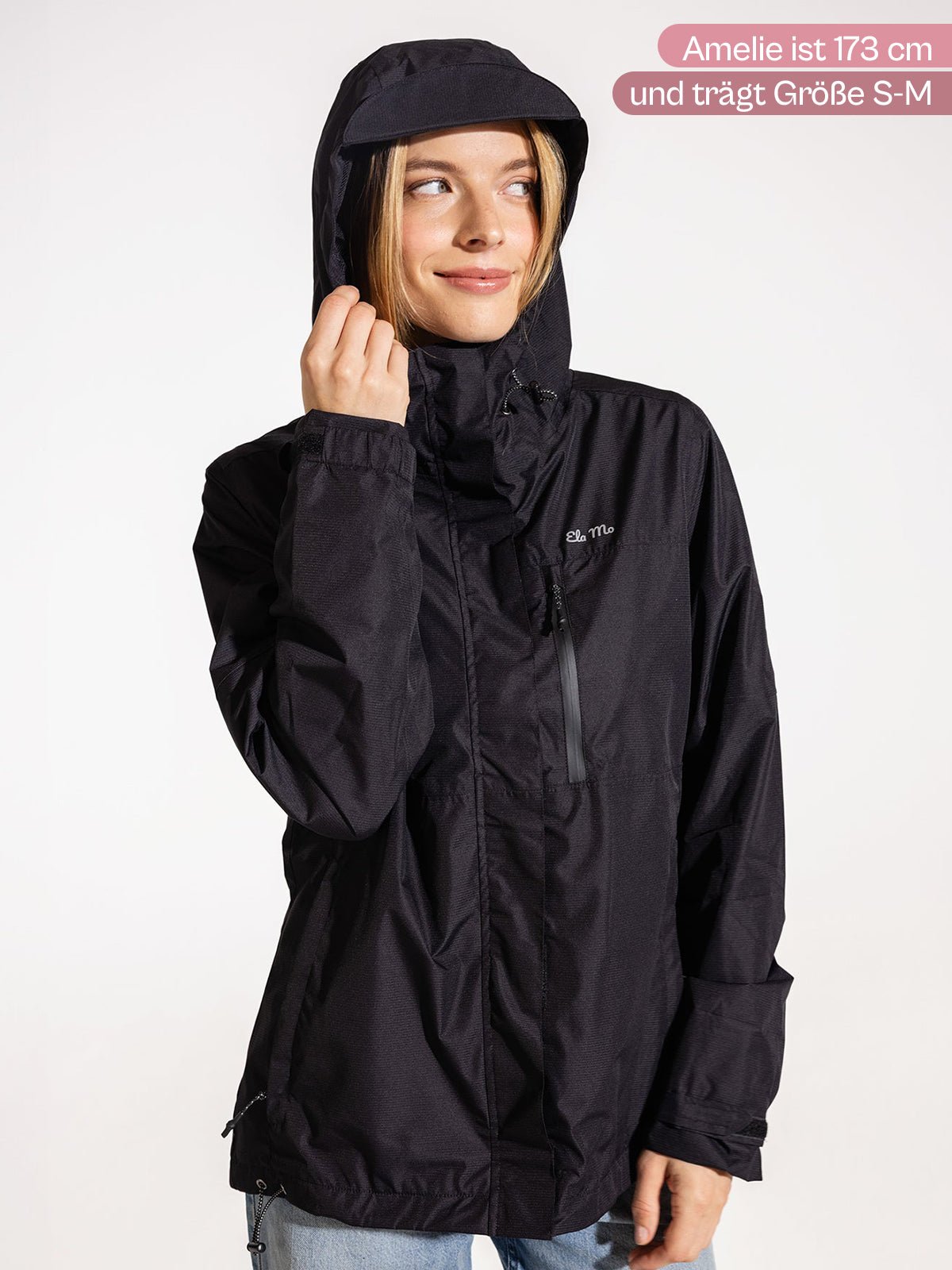 Ela Mo™ Regenjacke | Black. Eine Frau präsentiert die Regenjacke mit Größenangabe vor rosa Hintergrund.