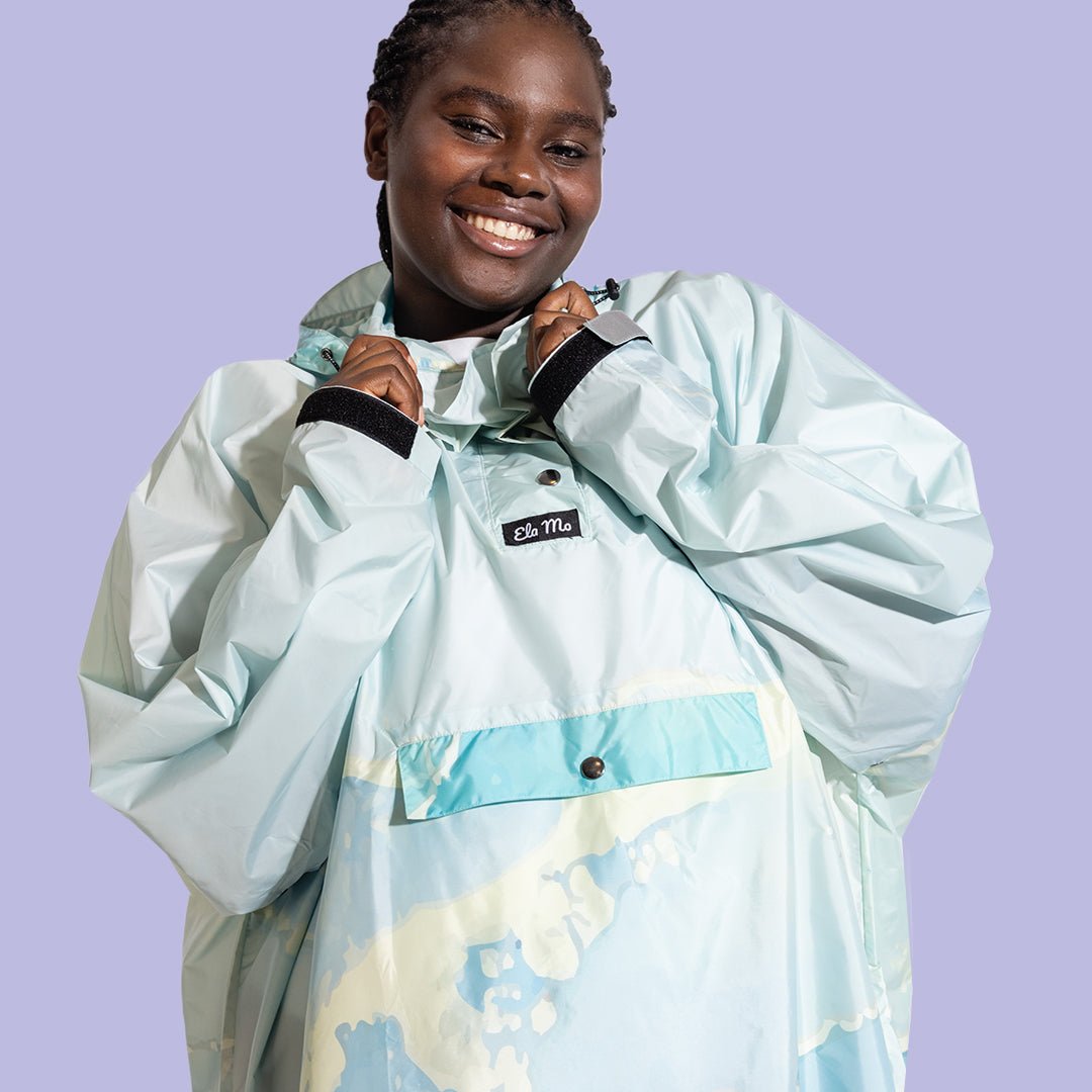 Ela Mo™ Regenponcho | Carribean Sea. Eine Frau in einem blauen Regenponcho vor lila Hintergrund.