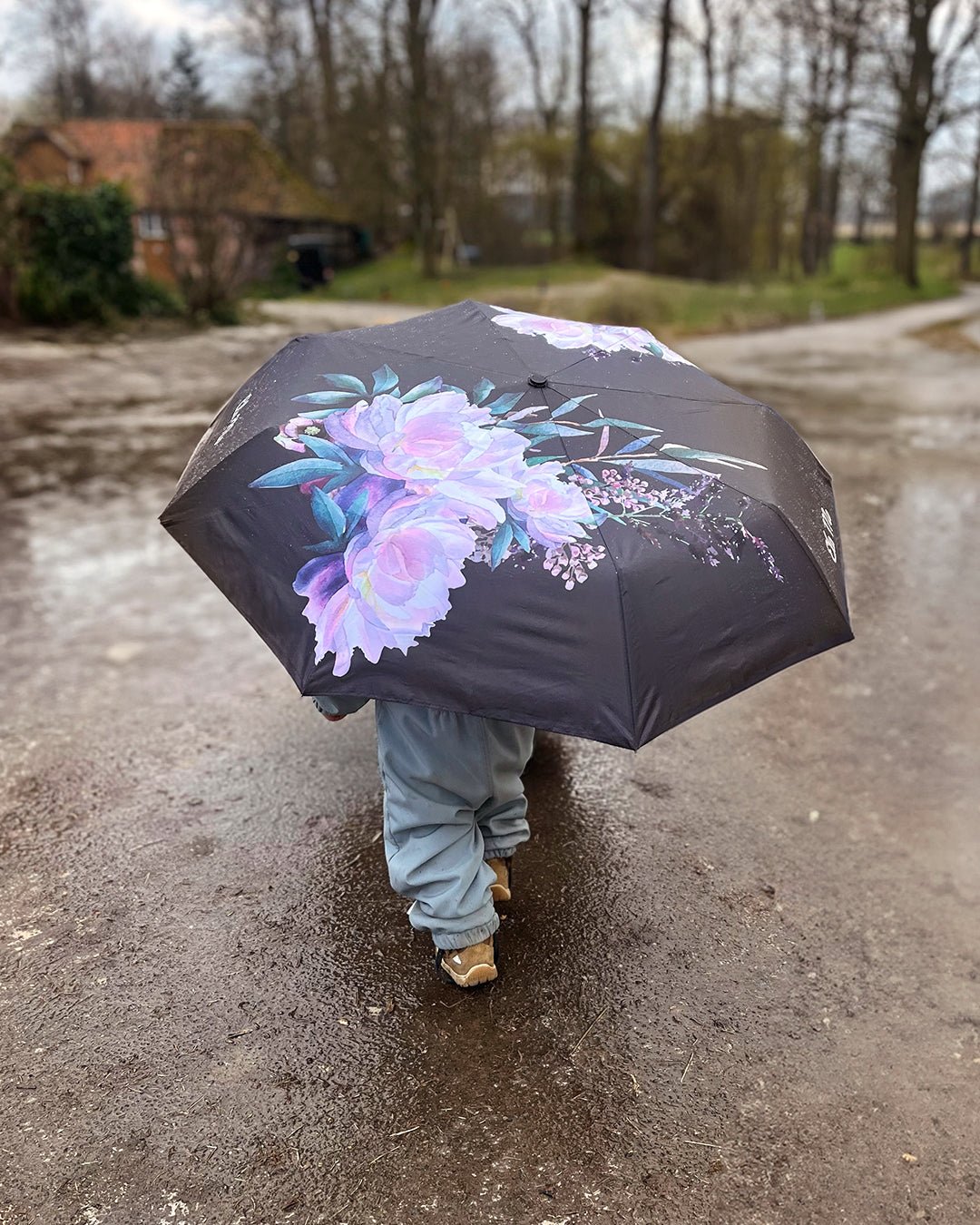 Ela Mo™ Taschenschirm | Beauty in Darkness. Kleines Kind mit schwarzem, geblümtem Schirm an einem Regentag.