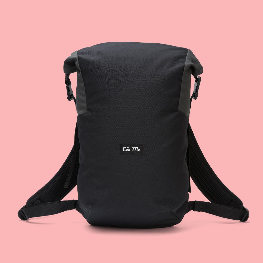 Ela Mo™ Rolltop Lite | Black. Ein schwarzer Rucksack mit Herstellerlogo vorne auf rosa Hintergrund.