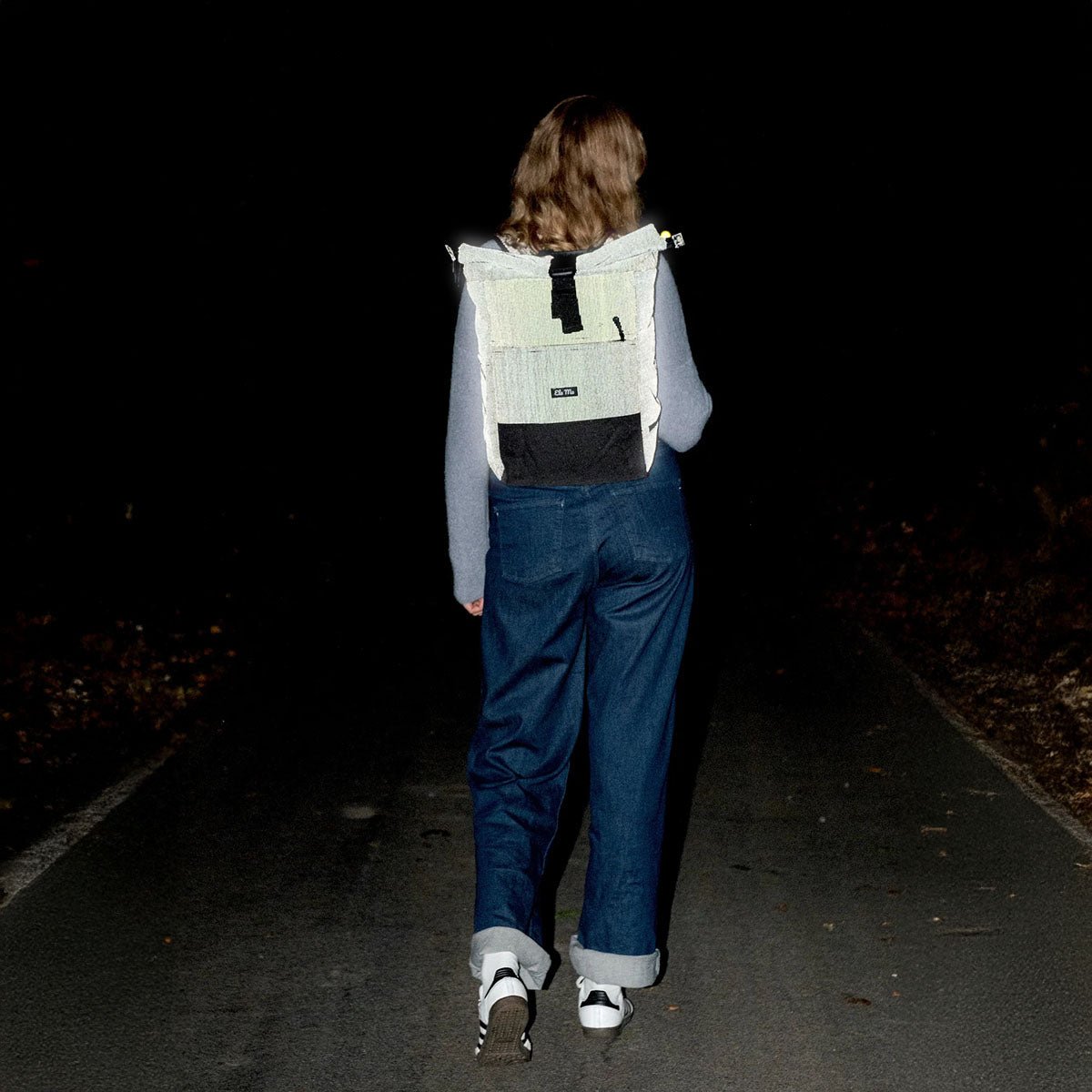 Ela Mo™ Rolltop Rucksack | Black Reflective. Eine Frau mit einem reflektierenden Rucksack in der Nacht.