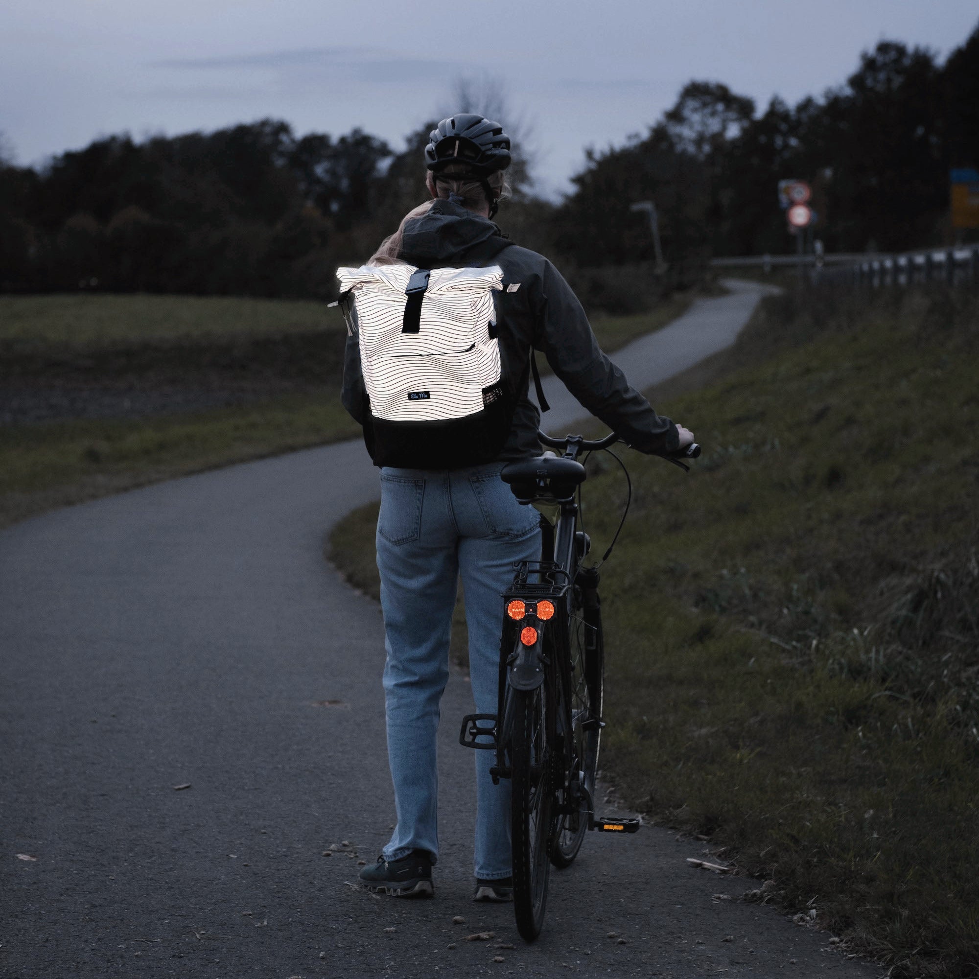 Ela Mo™ Rolltop Rucksack Night Edition | Wave. Frau mit reflektierendem Rucksack und Fahrrad auf der Wiese in der Nacht.
