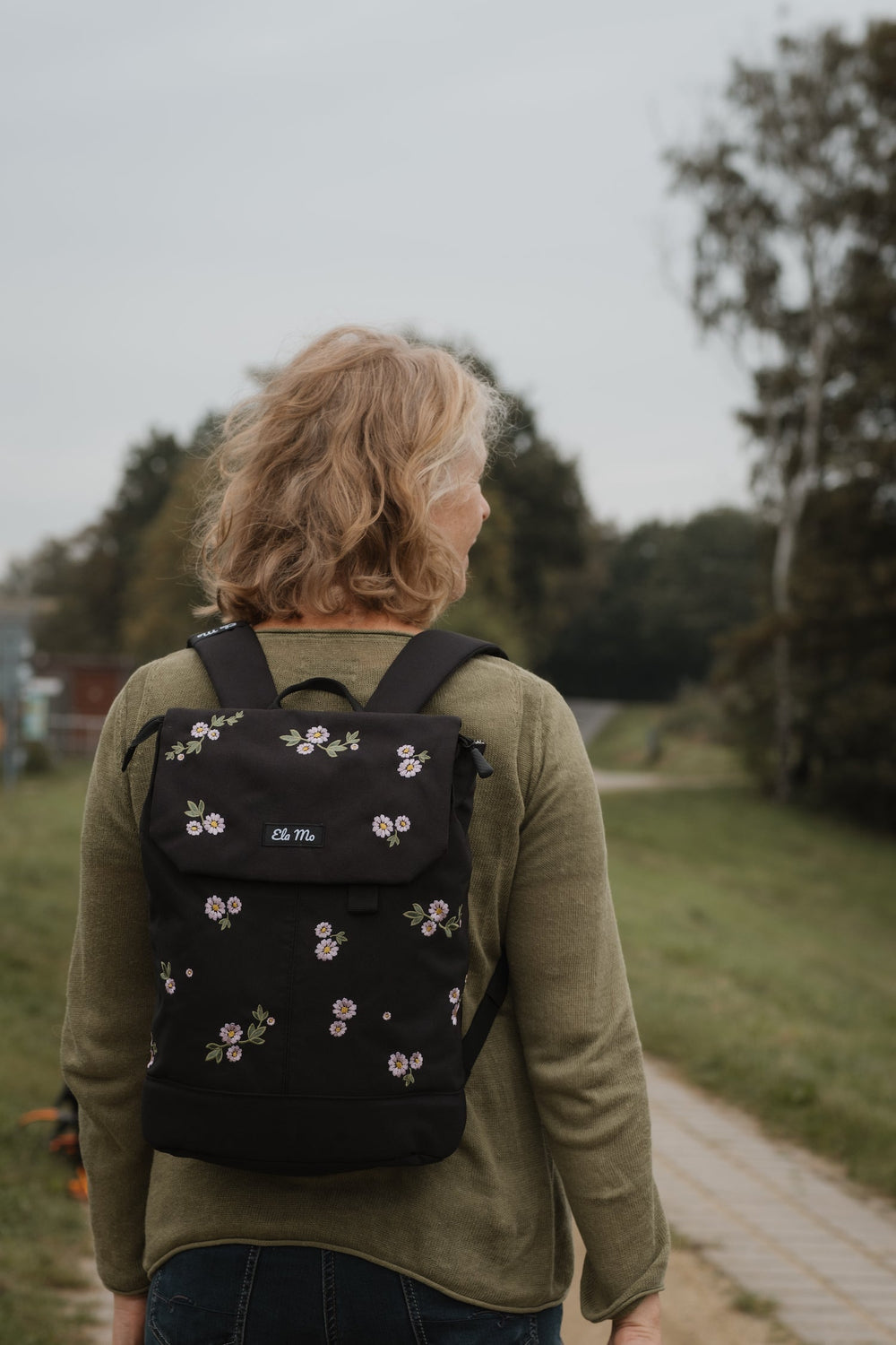 Ela Mo™ Daypack Rucksack | Embroidery Edition Upsy Daisy. Eine Frau mit dem Rucksack auf dem Rücken im Freien.