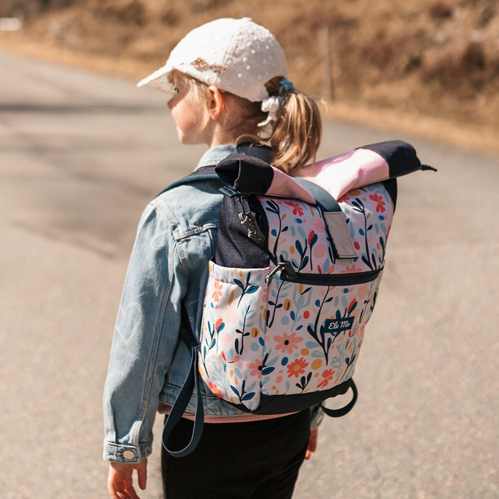 Ela Mo™ Kinderrucksack (6-10 Jahre) | Pastel Land. Ein kleines Mädchen trägt einen floral gemusterten Rucksack.