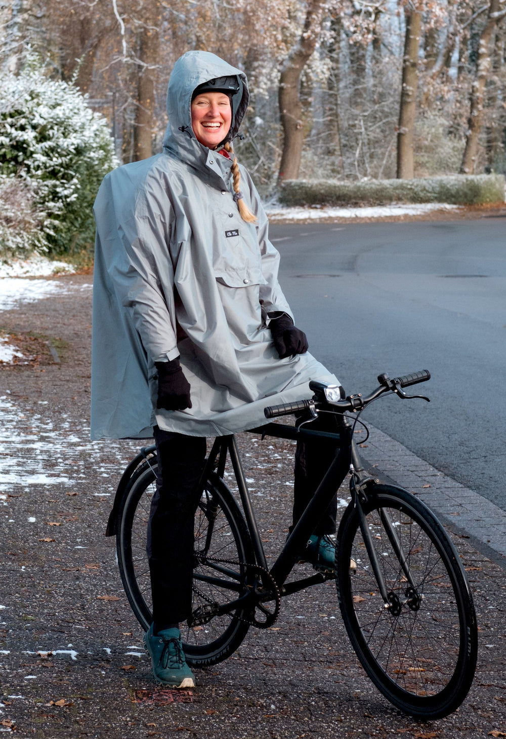 Ela Mo™ Regenponcho | Black. Eine Frau mit Regenponcho und Fahrrad auf der Straße.