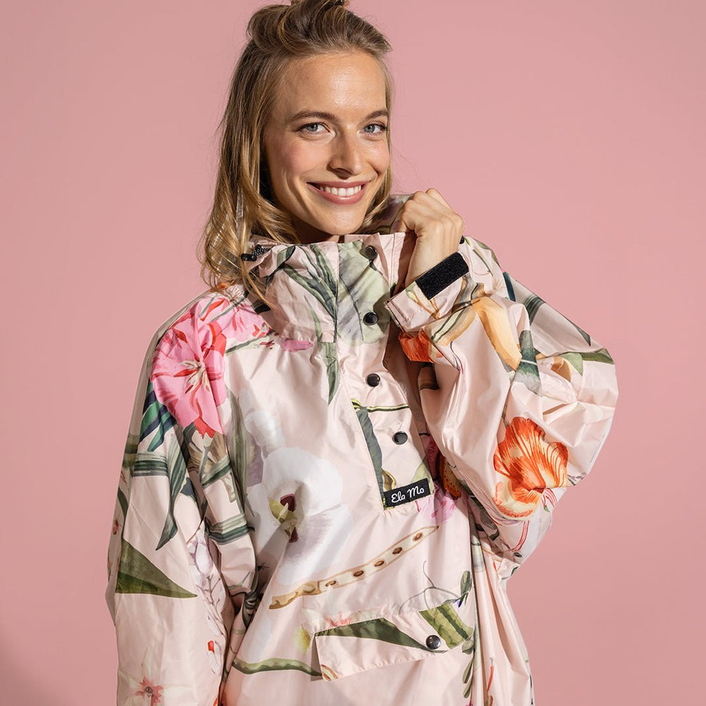 Ela Mo™ Regenponcho | Luxe. Eine Frau in einem floral gemusterten Regenponcho auf rosa Hintergrund.