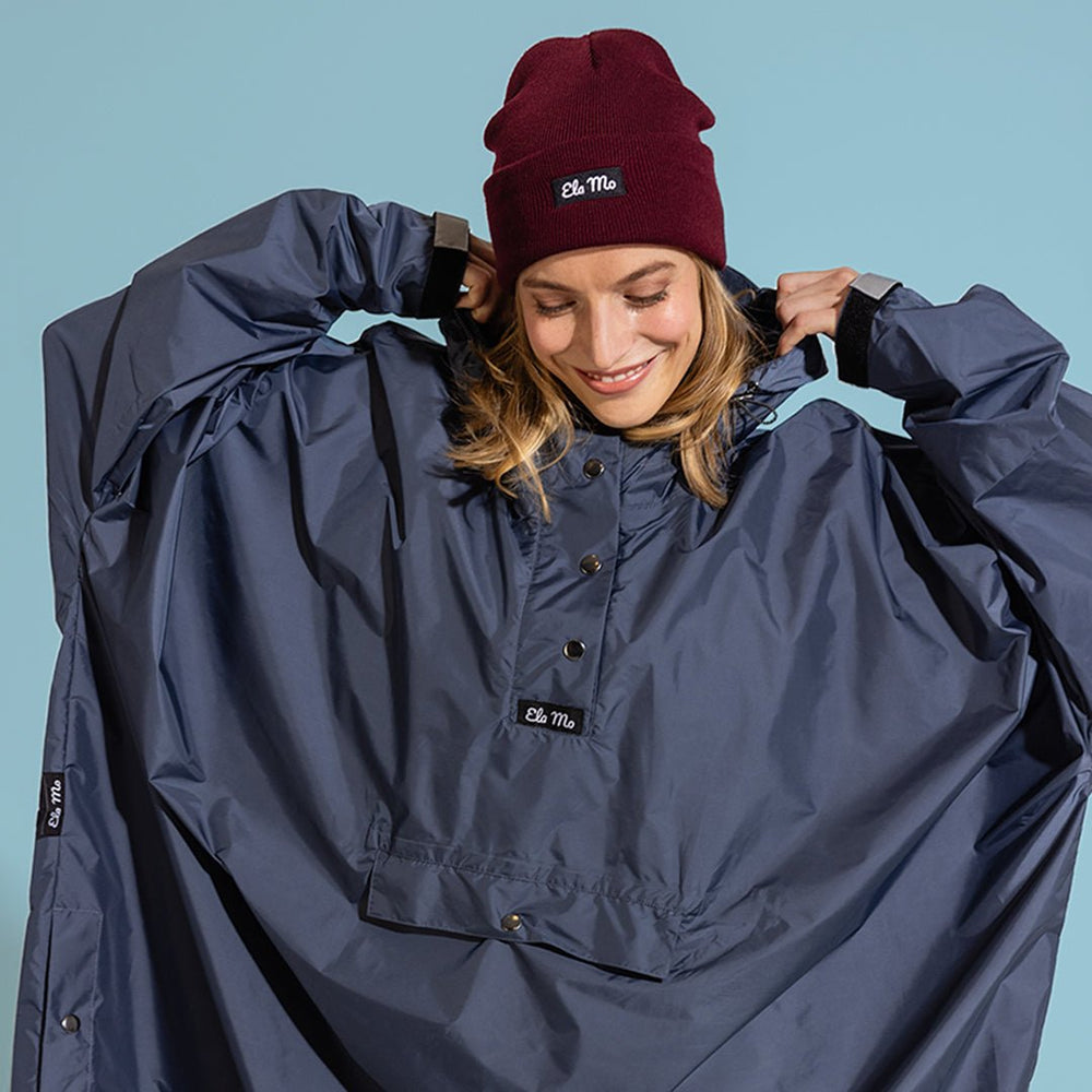 Ela Mo™ Regenponcho | Navy. Eine Frau in einem dunkelblauen Regenponcho auf blauem Hintergrund.