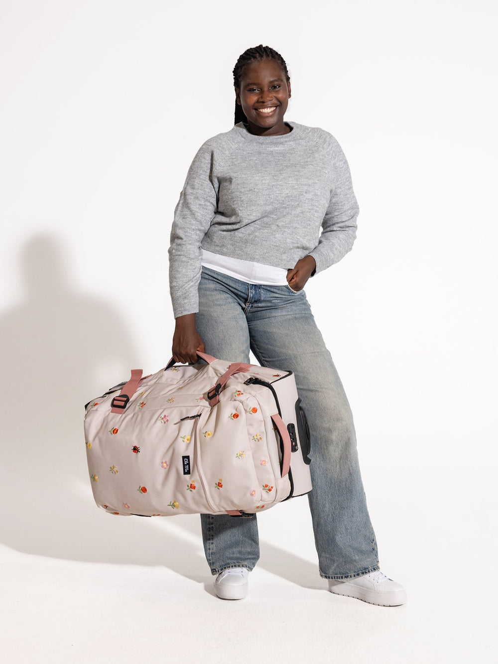 Ela Mo™ Reisetasche mit Rollen 78 Liter | Embroidery Edition Losy. Eine Frau hält die Tasche seitlich, Griff unten.